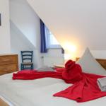 Schlafzimmer mit Dachschräge, Bett mit roter Decke, Holzkommode und blauer Stuhlecke.