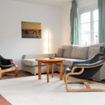 Wohnzimmer mit Sofa, zwei schwarzen Ledersessel und Holztisch. Fenster mit grünen Vorhängen.