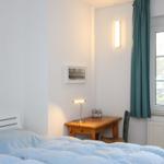 Schlafzimmer mit Bett, Tisch, Stuhl und Fenster. An der Wand hängt ein Bild und eine Lampe.