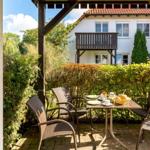 Terrasse mit Tisch und Stühlen, umgeben von grünen Hecken und einem weißen Haus im Hintergrund.