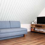 Ein Wohnzimmer mit blauem Sofa, Holzfußboden und schräger Holzwand. Fernseher auf Holzregal.