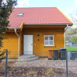 Holzhaus mit rotem Dach, weiße Tür und Fenster, Hof mit Zaun und Mülltonnen.