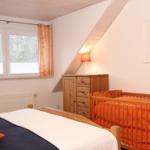 Schlafzimmer mit Babybett, Holzkommode und Fenster mit Blick auf Wald.