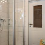 Dusche mit Glaswand, weiße Fliesen und Handbrause. Tür mit Glasfenster und Handtuch auf Badebank.