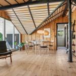 Ein Raum mit Holzdecke, -wänden und -boden. Es gibt einen Tisch mit Stühlen, einen Sessel und große Fenster.