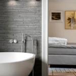 Bad mit freistehender Badewanne und grauer Wand. Im Hintergrund Schlafzimmer mit Bett und Kunstwerken.