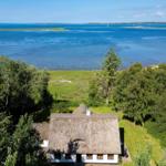 Haus mit Strohdach umgeben von Bäumen und Gras, Blick auf blaues Wasser und Insel.