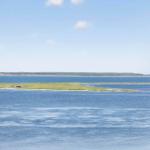 Weitläufiger Blick auf Wasserfläche mit Insel und Horizont unter blauem Himmel.