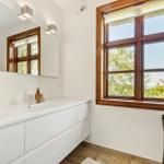 Badezimmer mit Waschbecken, Spiegel und Holzfenster mit Blick auf Bäume.