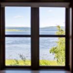 Durch das Fenster blickt man auf einen See mit grüner Ufervegetation und einem Wald im Hintergrund.