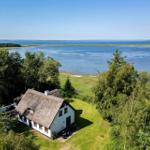 Haus mit Strohdach liegt am Wasser, umgeben von Grün und Bäumen.