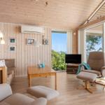 Wohnzimmer mit Holzwand, Sofa, Sessel und Fernseher. Große Fenster mit Blick auf Grün.