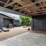 Terrasse mit Liegen, Grill und Tisch unter Holzkonstruktion. Schwarze Fassade und Steinplatten.