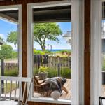 Durch die Fenster ist eine Terrasse mit Sitzgelegenheit und Blick auf Grün und Meer zu sehen.