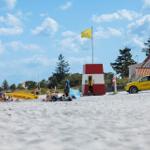Strand mit Rettungswagen, Leuchtturm und Menschen. Gelbe Flagge weht im Wind.