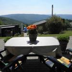 Terrasse mit Tisch, Stühlen und Blumen. Blick auf Hügel und Grünflächen.