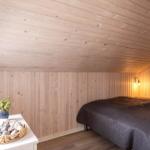 Schlafzimmer mit Holzwand und -decke, Bett mit dunkler Bettwäsche, Nachttisch mit Stein- und Pflanzenarrangement.