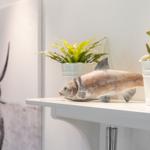 Ein Holzfigur eines Fisches steht auf einer weißen Ablagefläche neben zwei Pflanzen in weißen Töpfen.