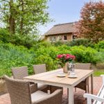 Terrasse mit Tisch, Stühlen und Blumen. Im Hintergrund ein Haus mit Schindeldach.