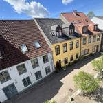 Haus mit gelber Fassade und rotem Dach in einer Straße mit gepflastertem Hof.