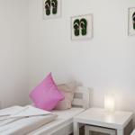 Zwei Betten mit Kissen und einer Nachttischlampe. An der Wand hängen drei Bilder mit Flip-Flops.
