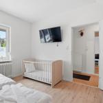 Zimmer mit Babybett, TV und Fenster. Tür führt in Wohnbereich.