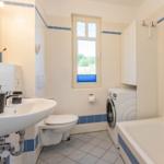 Badezimmer mit Waschmaschine, Badewanne und Toilette. Fenster mit blauen Jalousien.