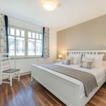 Schlafzimmer mit weißem Bett, Nachttischen und Holzboden. Fenster mit Vorhängen.