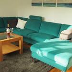 Ein Wohnzimmer mit einem türkisfarbenen Ecksofa, einem Holztisch und Wandbildern.