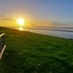 Sonnenuntergang über dem Wasser mit grünem Hang und Holzgeländer