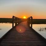 Holzbrücke führt durch ruhiges Wasser zu Sonnenuntergang über Grasland