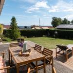 Terrasse mit Holztisch, Stühlen und Liegen. Grünfläche und Hecke im Hintergrund.