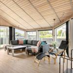 Ein Wohnbereich mit Holzboden, großem Sofa und vielen Fenstern. Draußen ist eine Terrasse mit Blick auf Grünflächen.