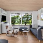 Wohnzimmer mit Kamin, Sofa und Sessel. Große Fenster bieten Blick auf Garten.