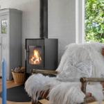 Ein Kaminofen mit brennendem Holz steht neben einem Stuhl mit Felldecke und Kissen.