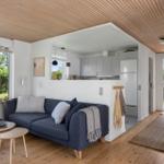 Wohnzimmer mit Sofa, Tischen und Blick auf Garten. Küche mit Kühlschrank sichtbar.