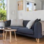 Ein Wohnzimmer mit grauem Sofa, zwei Holztischen und Blick auf Bäume.