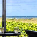 Durch das Fenster ist das Meer und Grünland zu sehen. Auf einem Tisch stehen zwei Gläser.