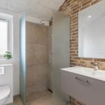 Bad mit Dusche, Waschbecken und Toilette. Wand aus Backstein und Fenster mit Pflanze.