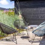Drei Stühle und ein Tisch auf einer Terrasse vor einem Holzhaus mit Gras und Blumen.