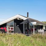 Schwarzes Holzhaus mit Terrasse, Sonnenschirm und Gartenmöbeln. Grün bewachsene Landschaft im Hintergrund.