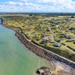 Aerial view of coastal village with houses on grassy dunes beside calm sea.