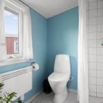 Badezimmer mit Toilette, Dusche und Fenster mit weißen Vorhängen.