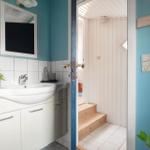 Badezimmer mit Waschbecken, Spiegel und Pflanze. Tür führt in einen Raum mit Holzstufen.