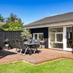 Terrasse mit Tisch, Stühlen und Grill. Haus mit Fenstern und Pflanzen.