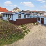 Blue holiday home with brown fence and wooden terrace. Sand path and green areas surround the accommodation.