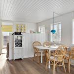 Dining room with round table, rattan chairs, and white wall paneling. Windows offer a view of the outside.