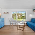 Ein Wohnzimmer mit blauem Sofa, grauer Sitzgelegenheit und Holzboden. Fenster zeigt Garten.