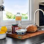 Küchenfenster mit Blick auf Garten, darauf Obst, Brot und Flasche.
