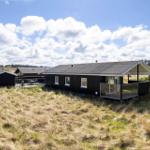 Modern wooden cabin with terrace in a grassy landscape under a blue sky with white clouds.